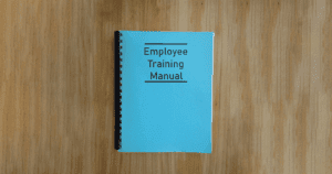 Blue Bound Book with Employee Training Manual on Cover Sitting on Light Wood Desk
