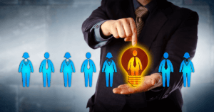 Line of Blue Businesspeople Icons in Front of Man in Suit Pointing to One Orange Man Icon in Lightbulb